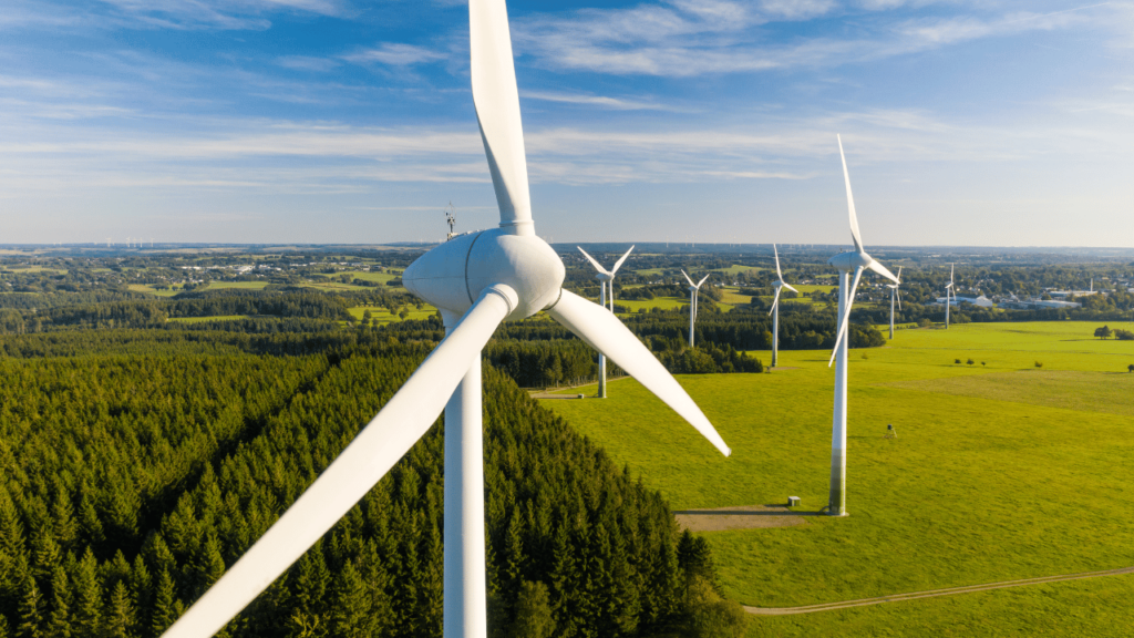 Aerial view of a wind farm with multiple wind turbines standing tall amidst green fields and forests under a clear blue sky, representing renewable energy initiatives in ESG (Environmental, Social, and Governance) Reporting Canada 2024.