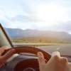 Driver's perspective from inside a car with hands on the steering wheel, embarking on a journey on an open road with a view of mountains in the distance. The scene evokes questions about practicalities of road trips, such as 'How Much Is Auto Insurance in Newfoundland and Labrador?