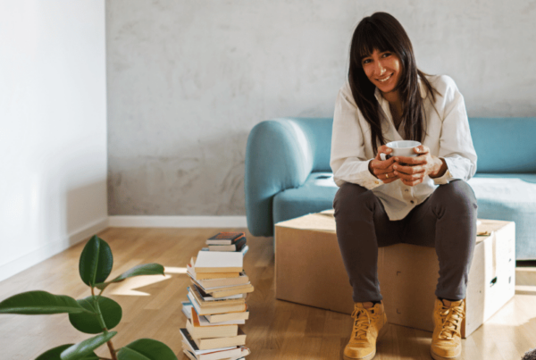 A content woman with a warm smile, holding a cup of coffee, is seated on a cardboard box in a sunlit living room, signifying a new move. Stacks of books lie by her side, with a plush blue sofa and a healthy green plant in the background. The cozy and minimalist decor emphasizes a fresh start, highlighting the importance of tenants insurance for new renters.