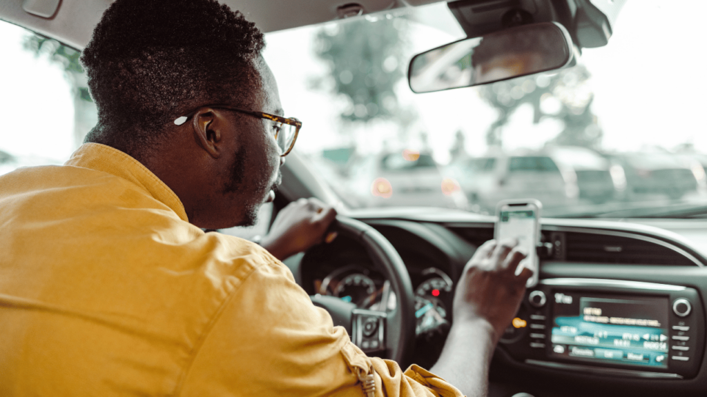 A person in a yellow shirt is driving a car, their left hand on the steering wheel and right hand holding a smartphone, possibly using a ride-sharing app like Uber, symbolizing Uber's arrival in Newfoundland and Labrador. The interior of the car and the view through the windshield suggest urban driving conditions.