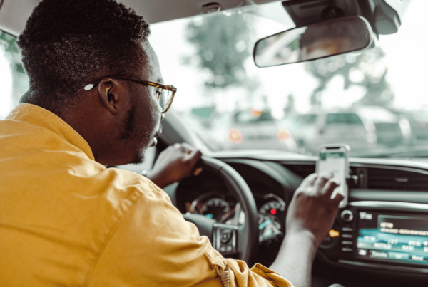 A person in a yellow shirt is driving a car, their left hand on the steering wheel and right hand holding a smartphone, possibly using a ride-sharing app like Uber, symbolizing Uber's arrival in Newfoundland and Labrador. The interior of the car and the view through the windshield suggest urban driving conditions.